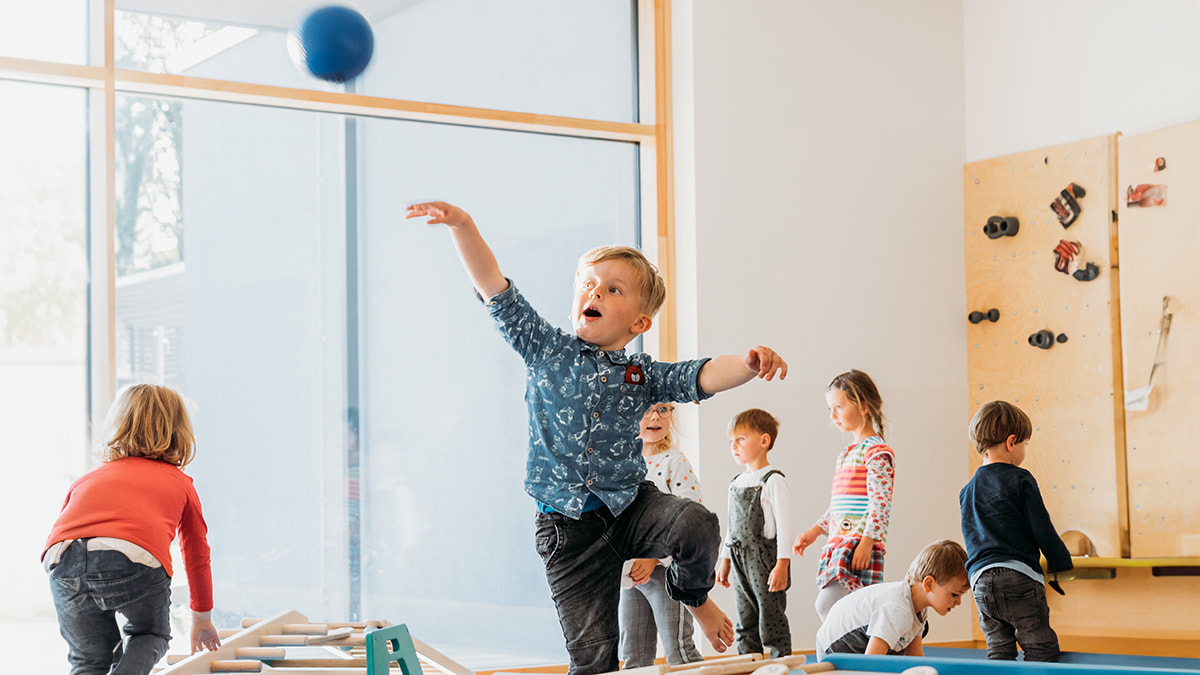  Junge wirft einen blauen Ball im Sportraum, während andere Kinder im Hintergrund spielen.