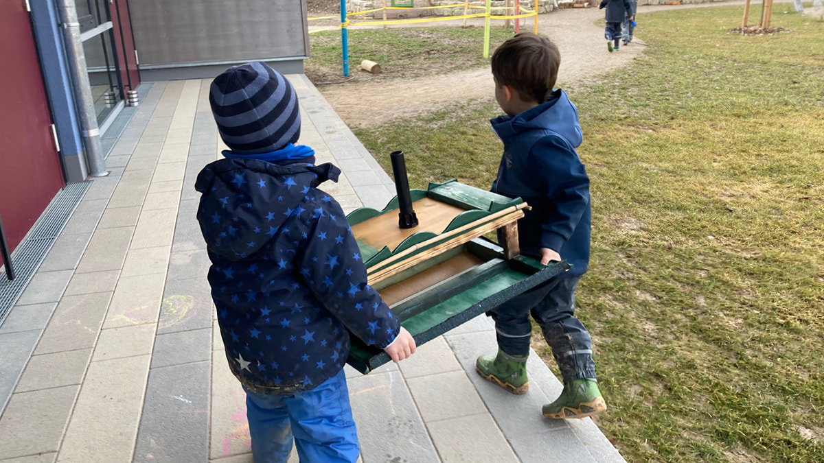 Zwei Kinder tragen gemeinsam ein großes Vogelhaus.