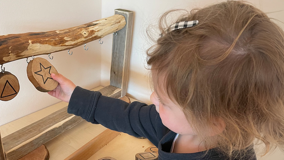 Kleinkind spielt mit hölzernen Formen, die an einem Gestell hängen.