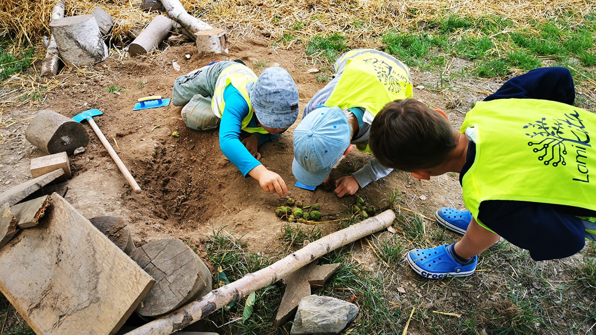  Drei Kinder in gelben Warnwesten spielen im Sand und sortieren grüne Eicheln, umgeben von Holzstücken und Werkzeug.