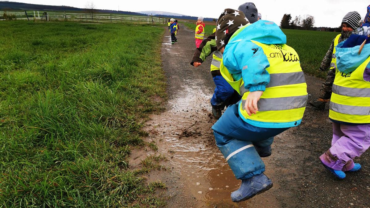 Kinder mit Warnwesten springen in eine schlammige Pfütze auf einem Feldweg.