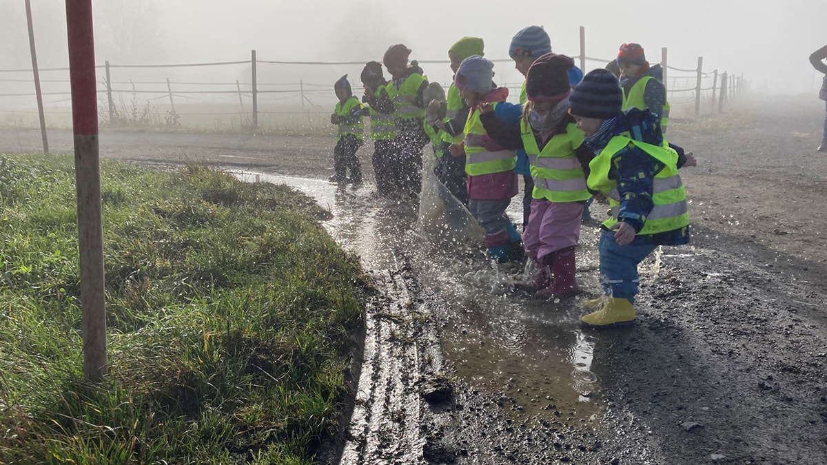 Eine Gruppe von LamiKita Kindern in Warnwesten und Gummistiefeln springt gemeinsam in eine Pfütze an einem nebligen Tag.