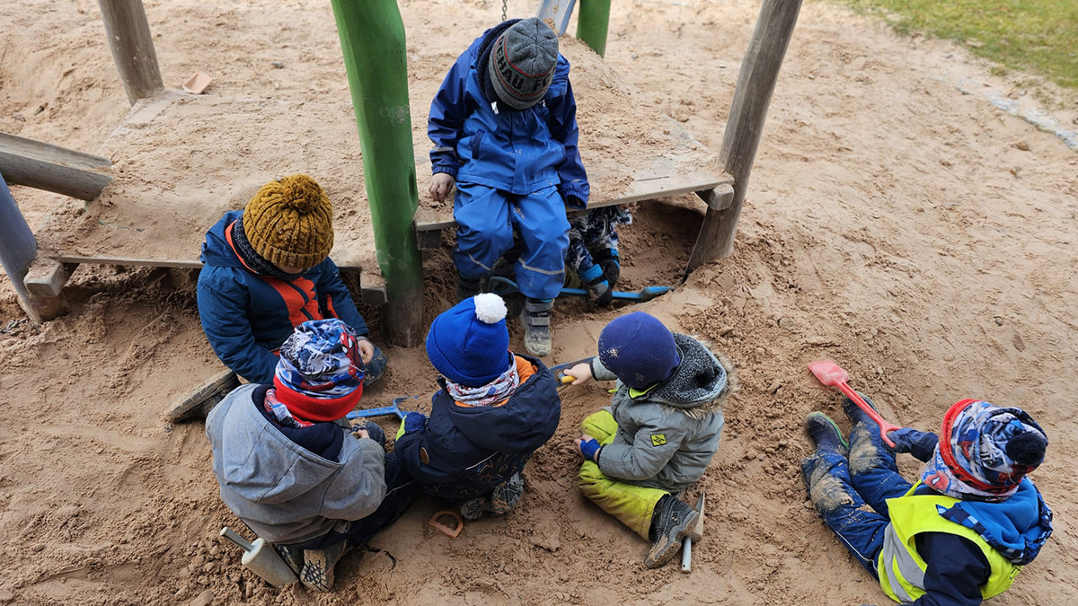 Kinder in Winterkleidung sitzen im Sandkasten und spielen mit Schaufeln und Werkzeugen.