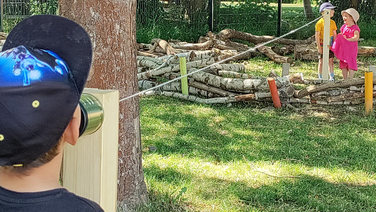 Kind spricht in ein Dosentelefon im Freien, während zwei andere Kinder am Ende der Schnur stehen und zuhören.