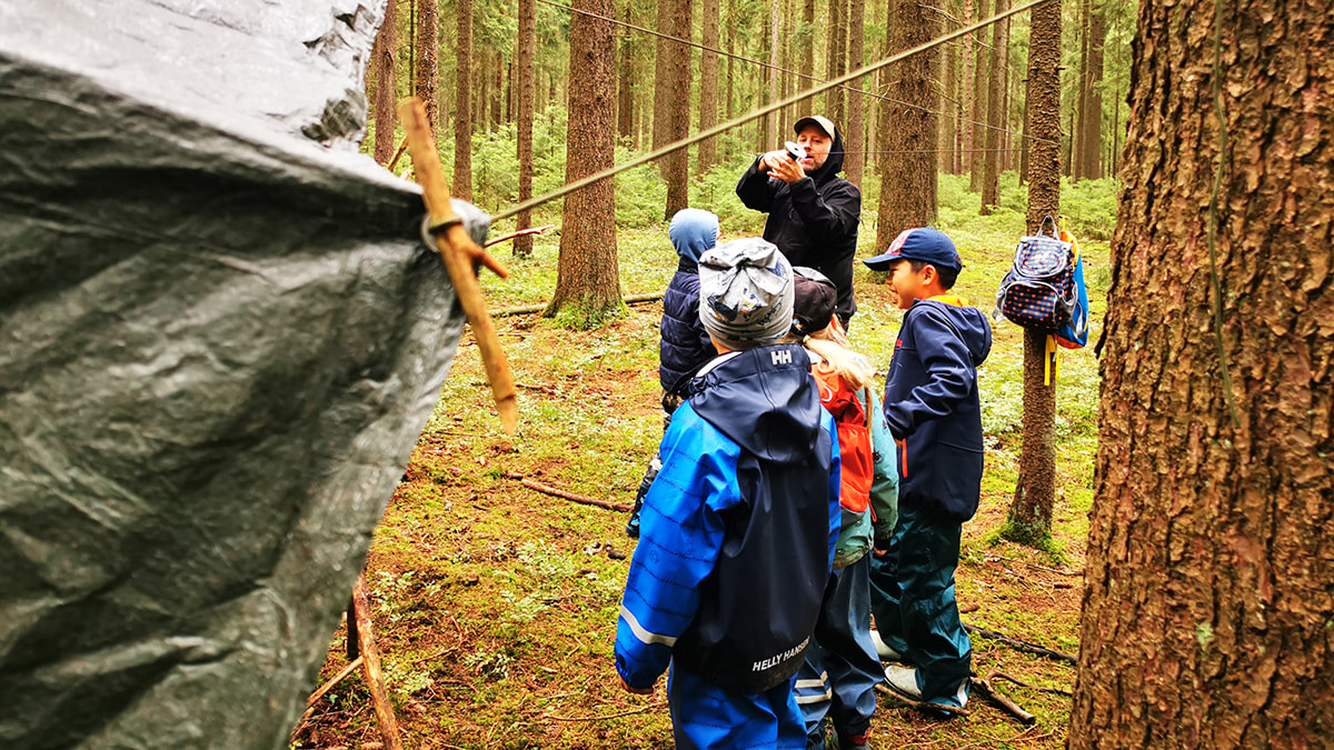 Gruppe von Kindern im Wald hört einem Erwachsenen zu, der vor einer aufgebauten Plane spricht.