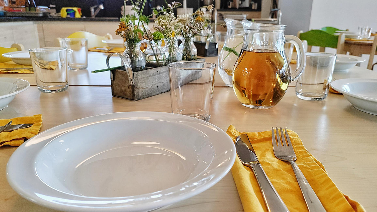 Gedeckter Esstisch mit weißen Tellern, gelben Servietten, Gläsern und einer Karaffe mit Tee, dekoriert mit Blumenvasen.